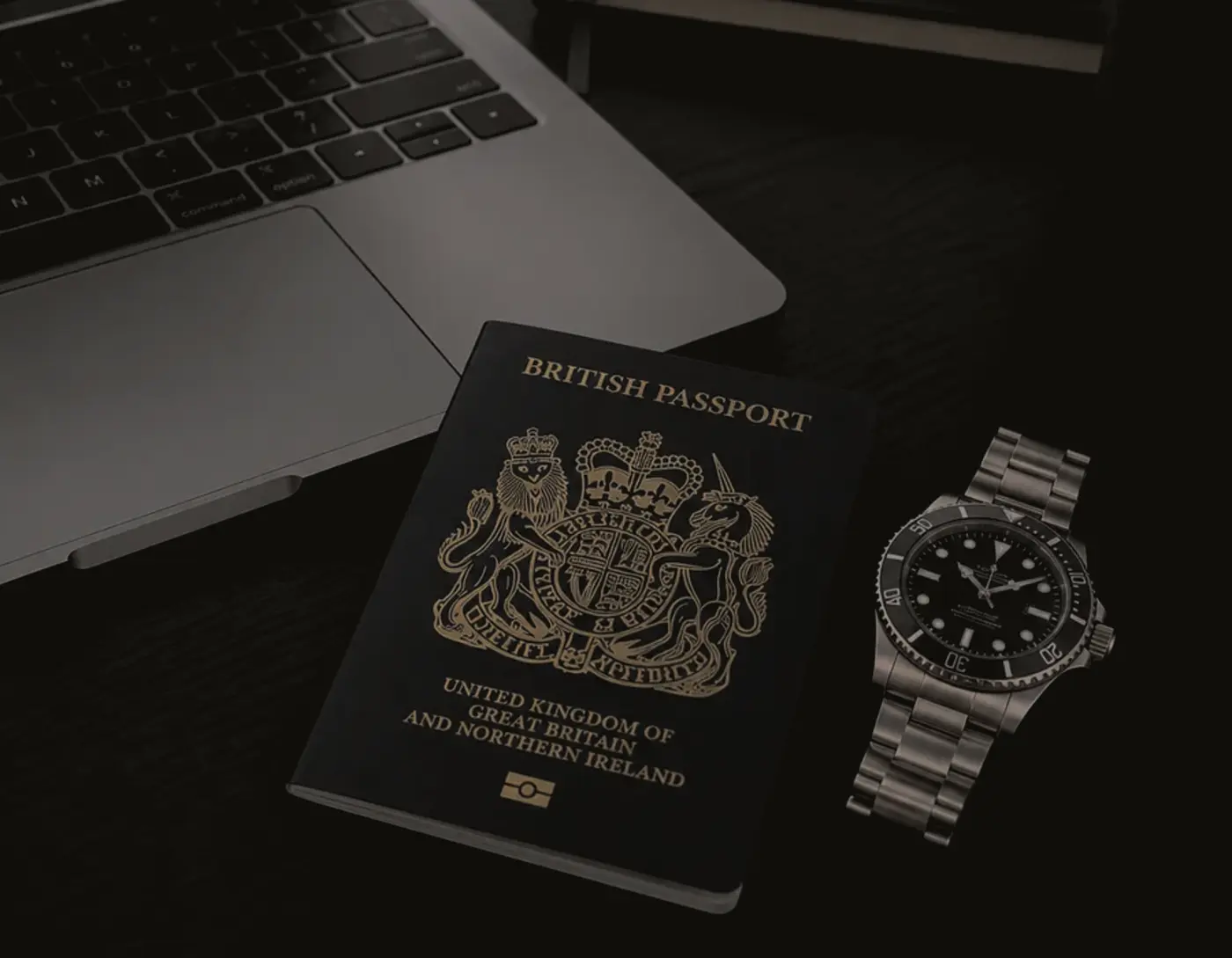 British passport and a wristwatch resting on a dark desk beside a laptop